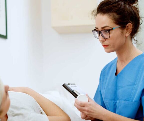 Close up of professional focused attractive nurse with eyeglasses sitting in front of woman patient with medications and clipboard at the hospital.