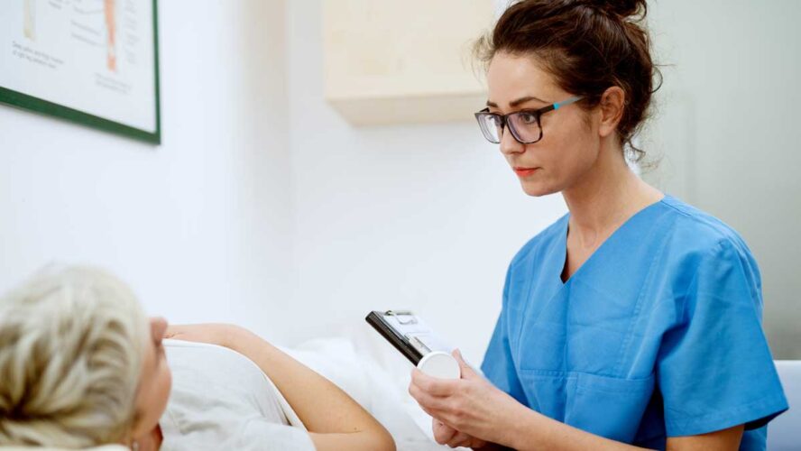 Close up of professional focused attractive nurse with eyeglasses sitting in front of woman patient with medications and clipboard at the hospital.