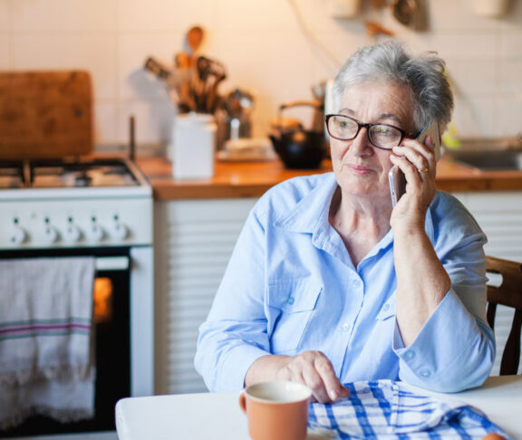 Senior woman is talking on mobile phone at cozy home kitchen. Grandmother has tea or coffee break in cooking. Retired person is using device. Concept of people cyber privacy, connection, communication