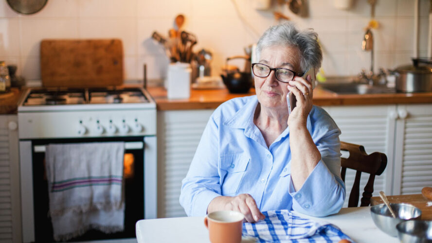 Senior woman is talking on mobile phone at cozy home kitchen. Grandmother has tea or coffee break in cooking. Retired person is using device. Concept of people cyber privacy, connection, communication