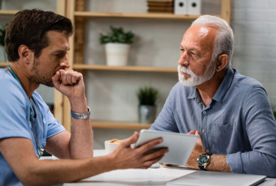 Senior man talking to a doctor who is using touchpad during medical appointment.