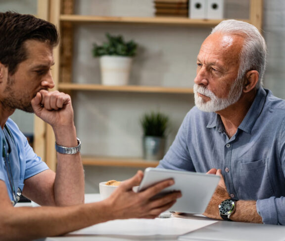 Senior man talking to a doctor who is using touchpad during medical appointment.