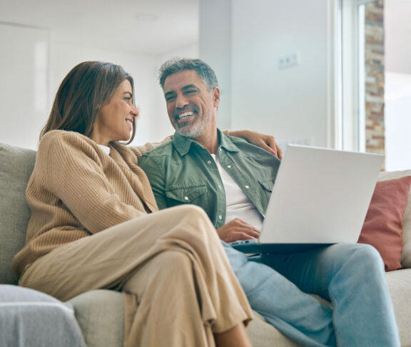 Happy middle aged couple using laptop computer relaxing on couch at home. Smiling mature man and woman talking having fun laughing with device sitting on sofa in sunny living room. Candid shot
