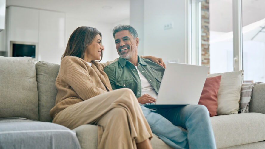 Happy middle aged couple using laptop computer relaxing on couch at home. Smiling mature man and woman talking having fun laughing with device sitting on sofa in sunny living room. Candid shot