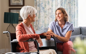 Mature woman comforting senior mom sitting on wheelchair at nursing home. Cheerful woman talking to old disabled mother in wheelchair at elder care centre. Loving caregiver taking care of elderly woman at home.