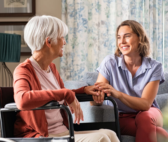Mature woman comforting senior mom sitting on wheelchair at nursing home. Cheerful woman talking to old disabled mother in wheelchair at elder care centre. Loving caregiver taking care of elderly woman at home.