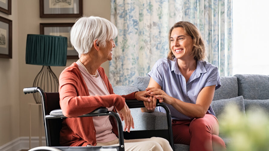 Mature woman comforting senior mom sitting on wheelchair at nursing home. Cheerful woman talking to old disabled mother in wheelchair at elder care centre. Loving caregiver taking care of elderly woman at home.