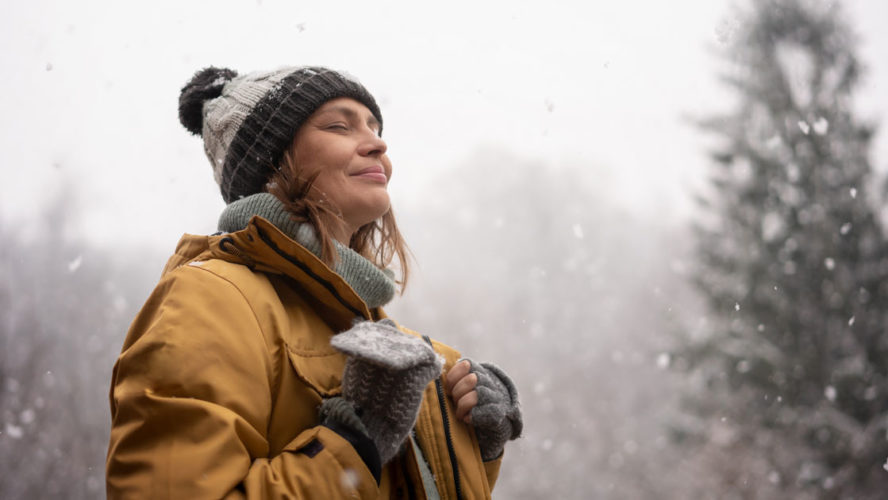 Young mature adult caucasian woman in a hat and yellow jacket breathing fresh air in the winter forest