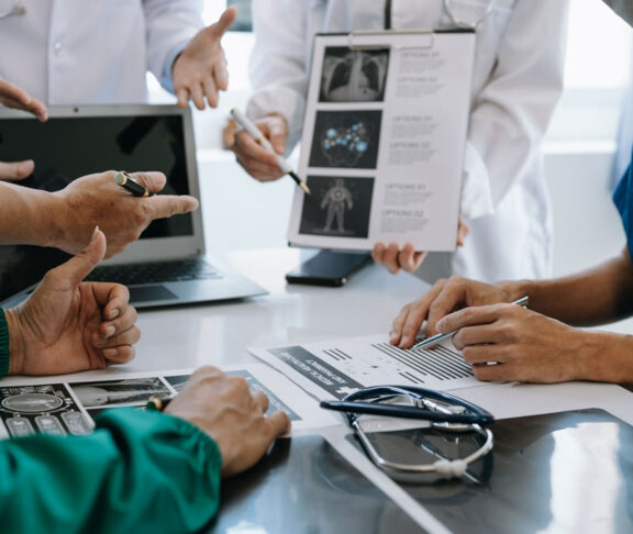 Medical team having a meeting with doctors in white lab coats and surgical scrubs seated at a table discussing a patients working online using computers in medical industry