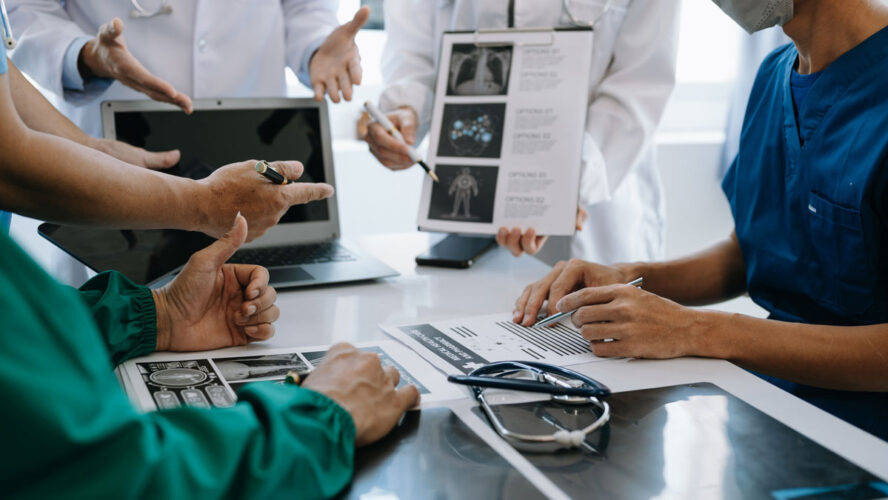 Medical team having a meeting with doctors in white lab coats and surgical scrubs seated at a table discussing a patients working online using computers in medical industry