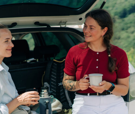 Two women friends are sitting in the back of car, talking and drinking coffee from a thermos during a trip out of city