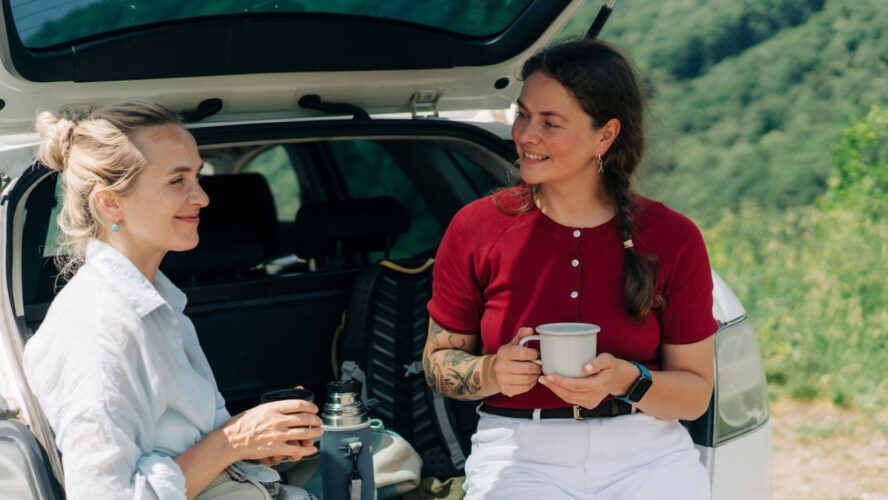 Two women friends are sitting in the back of car, talking and drinking coffee from a thermos during a trip out of city