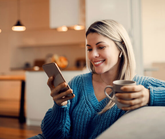 A happy woman hanging on social media on the phone and drinking coffee at home.