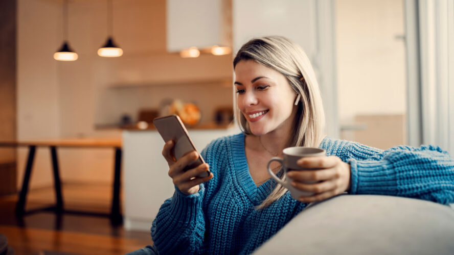 A happy woman hanging on social media on the phone and drinking coffee at home.
