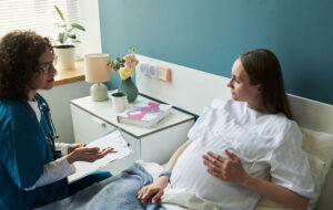 Caucasian pregnant young adult woman lying in hospital bed talking with Caucasian middle aged female doctor holding clipboard, medical consultation during pregnancy checkup