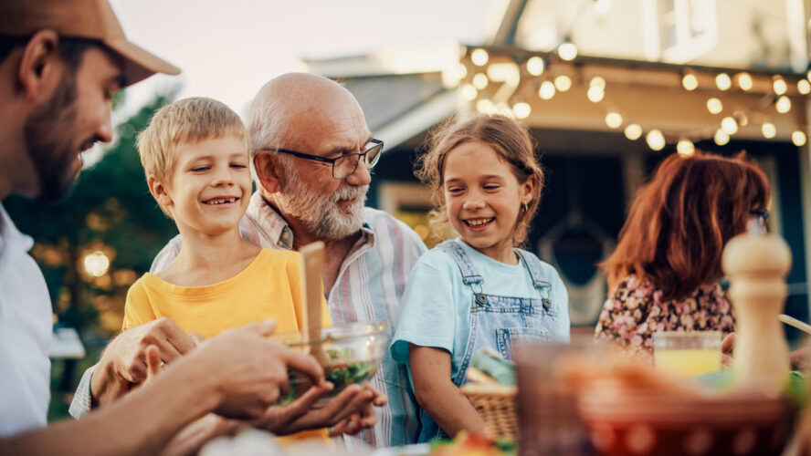 Happy Senior Grandfather Talking and Having Fun with His Grandchildren, Holding Them on Lap at a Outdoors Dinner with Food and Drinks. Adults at a Garden Party Together with Kids