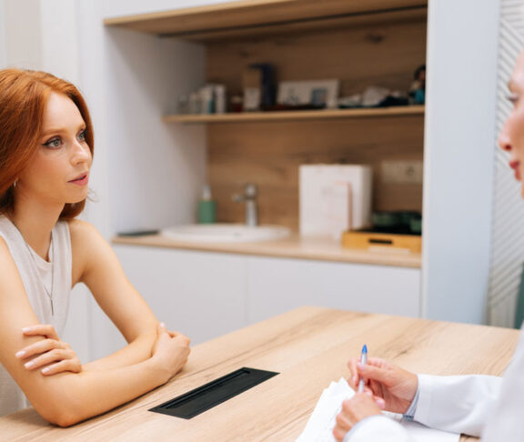 Side view of skilled middle-aged female doctor giving psychological support to young woman patient, speaking. Redhead lady visitor talking with practitioner for healthcare checkup in medical clinic