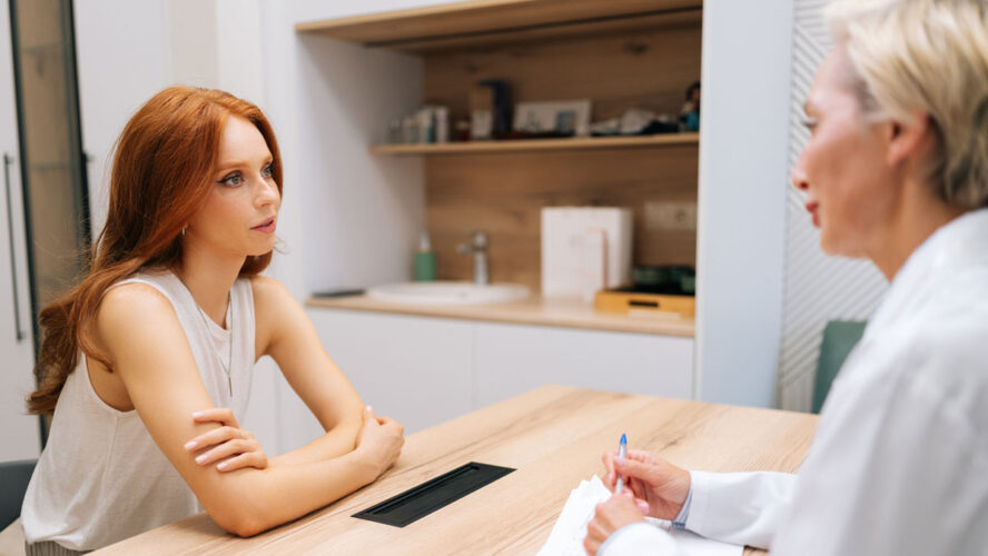 Side view of skilled middle-aged female doctor giving psychological support to young woman patient, speaking. Redhead lady visitor talking with practitioner for healthcare checkup in medical clinic