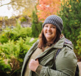 Young cheerful curvy woman in grey hat and green jacket walking in the park