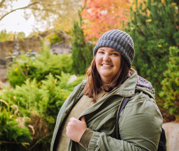 Young cheerful curvy woman in grey hat and green jacket walking in the park