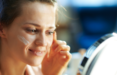 Elegant female applying facial cream and looking in mirror